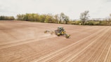 A tractor is working in a large, plowed agricultural field with trees in the background. The machinery is spreading over the field, preparing the soil for planting.