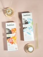 Two boxes of Bolsius True Moods diffusers are placed on a neutral pink surface. One box is labeled 'New Energy' with grapefruit and ginger scents featuring a vibrant orange and red design. The other is labeled 'In Balance' with white tea and mint leaves, displaying a cool blue and green design. Next to the boxes are reed diffusers in glass containers, with a set of bamboo sticks leaning against the surface.