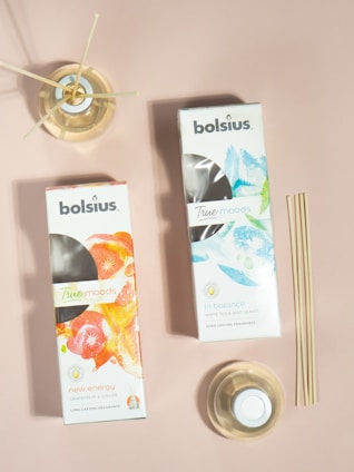 Two boxes of Bolsius True Moods diffusers are placed on a neutral pink surface. One box is labeled 'New Energy' with grapefruit and ginger scents featuring a vibrant orange and red design. The other is labeled 'In Balance' with white tea and mint leaves, displaying a cool blue and green design. Next to the boxes are reed diffusers in glass containers, with a set of bamboo sticks leaning against the surface.