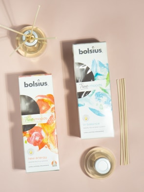 Two boxes of Bolsius True Moods diffusers are placed on a neutral pink surface. One box is labeled 'New Energy' with grapefruit and ginger scents featuring a vibrant orange and red design. The other is labeled 'In Balance' with white tea and mint leaves, displaying a cool blue and green design. Next to the boxes are reed diffusers in glass containers, with a set of bamboo sticks leaning against the surface.