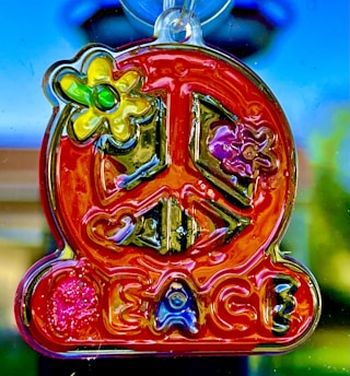 A colorful, translucent peace sign ornament with embedded flowers is hanging. The word 'Peace' is inscribed at the bottom. The vibrant setting includes red, yellow, and blue hues against a blurred outdoor background.