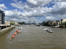 A river with boats and ships traversing it, flanked by buildings on both sides. The iconic structure of Tower Bridge stands in the background. The sky is partly cloudy, creating a mix of sunlight and overcast shadows.