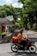 A person riding a motorcycle is captured in motion along a tree-lined urban street. The motorcycle is equipped with a large orange delivery box, and the rider is wearing a matching orange jacket and helmet. On the left, a covered sidewalk or pedestrian area is visible, and traffic cones are placed beside it. Tall trees with lush green foliage provide shade and a serene backdrop to the scene.