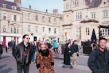 A bustling urban setting features several people walking, some wearing sunglasses and stylish clothing. The backdrop consists of historic stone buildings with ornate architecture, reflecting a blend of modern life and old-world charm. Bicycles and pedestrians share the space, conveying activity and movement.