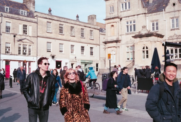 A bustling urban setting features several people walking, some wearing sunglasses and stylish clothing. The backdrop consists of historic stone buildings with ornate architecture, reflecting a blend of modern life and old-world charm. Bicycles and pedestrians share the space, conveying activity and movement.