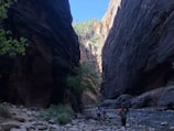 A dramatic canyon scene with steep, dark rock walls rising on either side. A narrow path runs alongside a shallow river, and several hikers are walking along the rocky trail. The sun illuminates the canyon peaks in the distance, highlighting lush greenery and contrasting the shadows below.