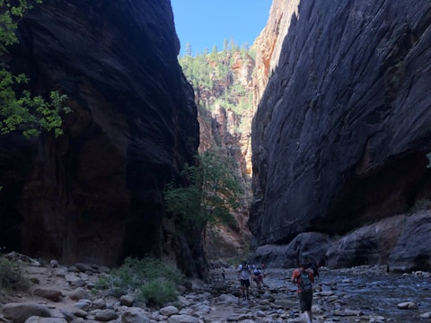 A dramatic canyon scene with steep, dark rock walls rising on either side. A narrow path runs alongside a shallow river, and several hikers are walking along the rocky trail. The sun illuminates the canyon peaks in the distance, highlighting lush greenery and contrasting the shadows below.