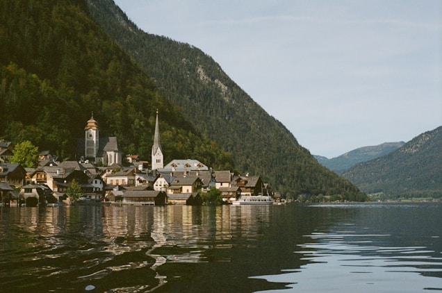 The image displays a picturesque lakeside village nestled at the foot of lush green mountains. There are several charming houses with grey roofs and a couple of prominent church steeples. The water in the foreground is calm, reflecting the buildings and the mountains in the background. The sky is lightly overcast with soft, diffused lighting.