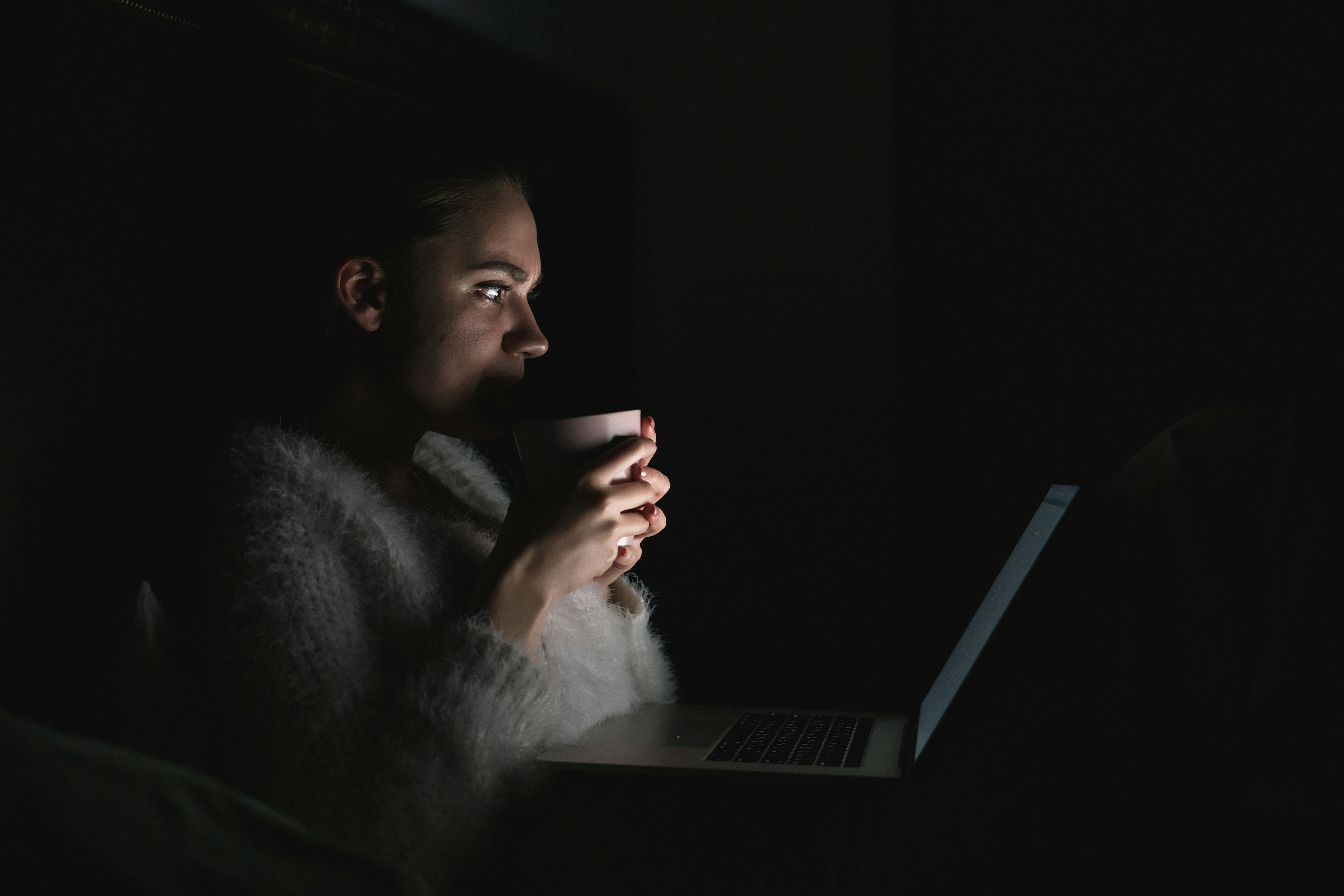 the girl lies in bed at night and carefully looks at the laptop screen. eating food, holding a mug and watching a series at night