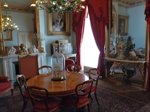 A luxurious interior featuring a round wooden table surrounded by elegant chairs with red upholstered seats. The room is adorned with opulent decorations, including a chandelier with green glass leaves, ornate picture frames, and intricate porcelain figurines. A large, gilded mirror with red curtains reflects part of the room, enhancing the grandeur. The walls are decorated with classic art pieces, and the overall decor suggests a historical or vintage theme.