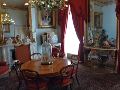 A luxurious interior featuring a round wooden table surrounded by elegant chairs with red upholstered seats. The room is adorned with opulent decorations, including a chandelier with green glass leaves, ornate picture frames, and intricate porcelain figurines. A large, gilded mirror with red curtains reflects part of the room, enhancing the grandeur. The walls are decorated with classic art pieces, and the overall decor suggests a historical or vintage theme.