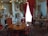 A luxurious interior featuring a round wooden table surrounded by elegant chairs with red upholstered seats. The room is adorned with opulent decorations, including a chandelier with green glass leaves, ornate picture frames, and intricate porcelain figurines. A large, gilded mirror with red curtains reflects part of the room, enhancing the grandeur. The walls are decorated with classic art pieces, and the overall decor suggests a historical or vintage theme.