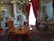 A luxurious interior featuring a round wooden table surrounded by elegant chairs with red upholstered seats. The room is adorned with opulent decorations, including a chandelier with green glass leaves, ornate picture frames, and intricate porcelain figurines. A large, gilded mirror with red curtains reflects part of the room, enhancing the grandeur. The walls are decorated with classic art pieces, and the overall decor suggests a historical or vintage theme.
