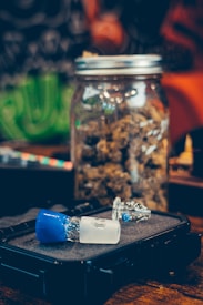 A jar filled with dried herbs or plant material sits on a wooden surface. In front of it, a black case holds a glass apparatus with a blue and frosted white piece. The background has blurred patterns and colors, suggesting an indoor setting.