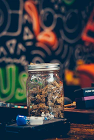 A mason jar filled with dried herbs sits on a table surrounded by other items. The background features a colorful, abstract mural with swirling patterns and vibrant colors.