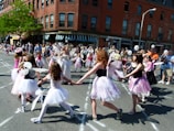 A group of children dressed as fairies hold hands in a circle on a city street during a parade or festival, while onlookers watch. The children wear wings and tutu-like skirts, and the surrounding crowd includes adults taking photos and supervising. The setting has a festive and lively atmosphere, with people enjoying a sunny day.