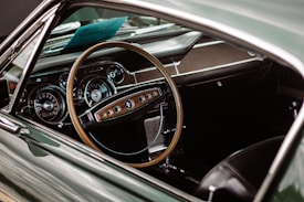 The image focuses on the interior of a vintage car, showcasing a wooden steering wheel with chrome accents and a dashboard featuring classic speedometers and gauges. The interior design reflects a classic, retro aesthetic with elements of dark leather and polished metal.