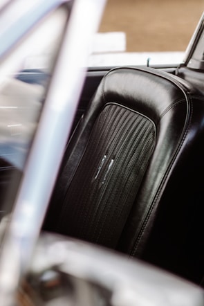 A close-up view of a car seat with black leather upholstery featuring detailed stitching and a textured center section. The surrounding frame of the car's interior is partially visible, with a soft out-of-focus effect.