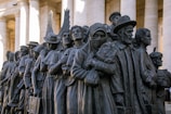 A bronze sculpture depicting a diverse group of people standing closely together. They appear to be of different cultural backgrounds, dressed in various historical and traditional clothing styles, and some hold personal belongings or wear headscarves.