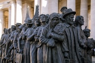 A bronze sculpture depicting a diverse group of people standing closely together. They appear to be of different cultural backgrounds, dressed in various historical and traditional clothing styles, and some hold personal belongings or wear headscarves.