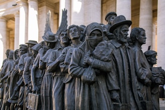 A bronze sculpture depicting a diverse group of people standing closely together. They appear to be of different cultural backgrounds, dressed in various historical and traditional clothing styles, and some hold personal belongings or wear headscarves.