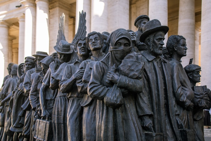 A bronze sculpture depicting a diverse group of people standing closely together. They appear to be of different cultural backgrounds, dressed in various historical and traditional clothing styles, and some hold personal belongings or wear headscarves.