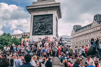 A large group of people gathered around a monument in a bustling public square. The monument features intricate carvings, and there are buildings with classical architecture in the background. People appear to be socializing and taking photos, and some are holding flags.