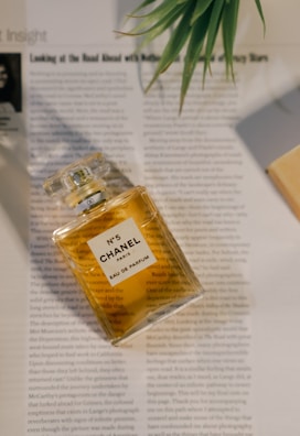 A bottle of Chanel No. 5 Eau de Parfum is placed on top of an open magazine, with a leafy green plant in the top right corner providing a touch of nature. The golden hue of the perfume contrasts with the black and white text on the pages.