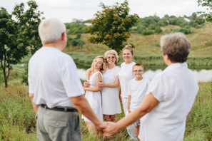 A family gathers outdoors on a grassy area with a scenic backdrop of a pond and trees. They are dressed in white, with two adults holding hands in the foreground, looking at three women and a child who are smiling at the camera. The setting appears calm and natural, with rolling hills in the distance.