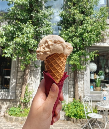 A hand is holding an ice cream cone with a scoop of beige ice cream in front of a blurred outdoor backdrop featuring greenery and a stone building with windows. The ice cream cone is wrapped with a dark red napkin, and there is a small cobblestone patio with white metal furniture visible in the background.