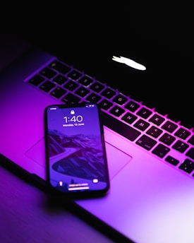 A smartphone displaying a lock screen with the time 1:40 and date Monday, 13 June rests on the keyboard of an open laptop. The scene is illuminated with a purple hue, giving it a modern and technological feel. The laptop's logo glows on the screen.