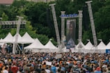A large crowd gathers outdoors in front of a stage and a large video screen displaying a person speaking into a microphone. White tents and tall metal scaffolding support structures can be seen around the stage. The surrounding environment is lush with green trees, creating a backdrop for the event. People in the crowd are wearing various colors, with many sporting blue shirts.