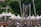 A large crowd gathers outdoors in front of a stage and a large video screen displaying a person speaking into a microphone. White tents and tall metal scaffolding support structures can be seen around the stage. The surrounding environment is lush with green trees, creating a backdrop for the event. People in the crowd are wearing various colors, with many sporting blue shirts.
