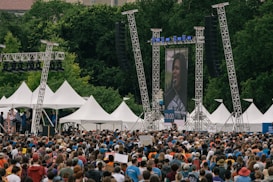 A large crowd gathers outdoors in front of a stage and a large video screen displaying a person speaking into a microphone. White tents and tall metal scaffolding support structures can be seen around the stage. The surrounding environment is lush with green trees, creating a backdrop for the event. People in the crowd are wearing various colors, with many sporting blue shirts.