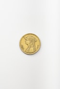 A single gold coin is placed on a plain, light-colored surface. The coin features an engraved image of a person with detailed hair styling, dressed in traditional garb. Intricate symbols and writing surround the portrait along the coin's edge.