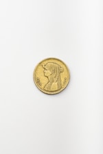A single gold coin is placed on a plain, light-colored surface. The coin features an engraved image of a person with detailed hair styling, dressed in traditional garb. Intricate symbols and writing surround the portrait along the coin's edge.