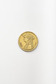 A single gold coin is placed on a plain, light-colored surface. The coin features an engraved image of a person with detailed hair styling, dressed in traditional garb. Intricate symbols and writing surround the portrait along the coin's edge.