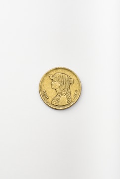 A single gold coin is placed on a plain, light-colored surface. The coin features an engraved image of a person with detailed hair styling, dressed in traditional garb. Intricate symbols and writing surround the portrait along the coin's edge.