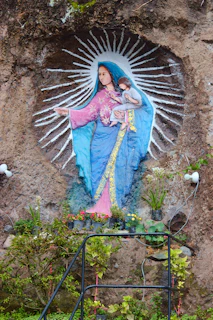 A painted religious mural featuring a figure in a blue robe with a pink dress, holding a child. The figure is surrounded by white radiating lines. Below the mural, there are potted plants and greenery, and parts of a metal railing are visible.