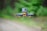 A compact drone equipped with a camera hovers in mid-air against a blurred green and brown outdoor background, highlighting its design and components.