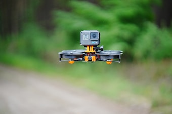 A compact drone equipped with a camera hovers in mid-air against a blurred green and brown outdoor background, highlighting its design and components.