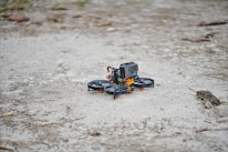 A small drone equipped with a GoPro camera is stationed on a rough and rocky ground. The drone has four propellers and is primarily black with orange accents.