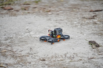 A small drone equipped with a GoPro camera is stationed on a rough and rocky ground. The drone has four propellers and is primarily black with orange accents.
