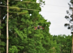 A drone with red propellers hovers mid-air against a backdrop of lush green trees. The landscape is peaceful, with overhanging power lines and blurred foliage creating depth.