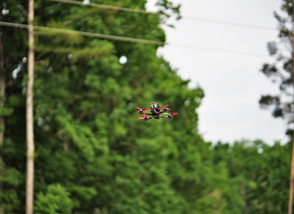 A drone with red propellers hovers mid-air against a backdrop of lush green trees. The landscape is peaceful, with overhanging power lines and blurred foliage creating depth.