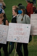 A group of people are gathered, holding signs with messages advocating for school safety and emphasizing the importance of learning over lockdowns. The signs are decorated with colorful messages and signatures. One person wearing a blue mask has a braid and wears a cap. The background shows a grassy area with more people.