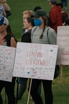 A group of people are gathered, holding signs with messages advocating for school safety and emphasizing the importance of learning over lockdowns. The signs are decorated with colorful messages and signatures. One person wearing a blue mask has a braid and wears a cap. The background shows a grassy area with more people.