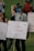 A group of people are gathered, holding signs with messages advocating for school safety and emphasizing the importance of learning over lockdowns. The signs are decorated with colorful messages and signatures. One person wearing a blue mask has a braid and wears a cap. The background shows a grassy area with more people.