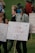 A group of people are gathered, holding signs with messages advocating for school safety and emphasizing the importance of learning over lockdowns. The signs are decorated with colorful messages and signatures. One person wearing a blue mask has a braid and wears a cap. The background shows a grassy area with more people.