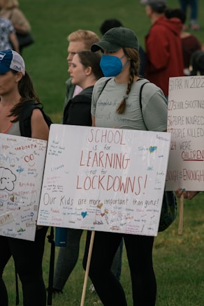A group of people are gathered, holding signs with messages advocating for school safety and emphasizing the importance of learning over lockdowns. The signs are decorated with colorful messages and signatures. One person wearing a blue mask has a braid and wears a cap. The background shows a grassy area with more people.
